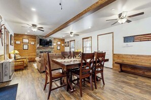 A dining area with a wooden table and chairs, a ceiling fan, and a television in the background. - Sparrow Cabin - East Village (Shaver Lake)