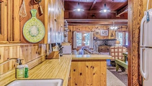 A wooden cabin interior with a kitchen area, wooden cabinets, a stone fireplace, and a dining table. - Alpine Cottage - West Village (Shaver Lake)