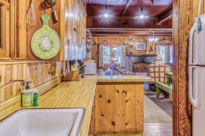 A wooden cabin interior with a kitchen area, wooden cabinets, a stone fireplace, and a dining table. - Alpine Cottage - West Village (Shaver Lake)