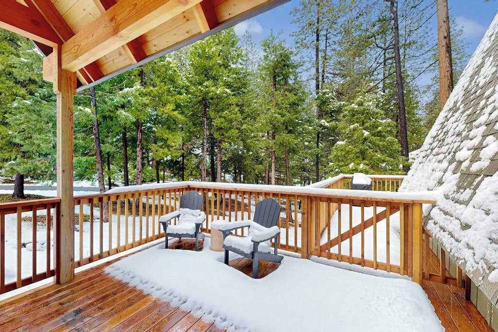 Una terraza de madera con dos sillas, rodeada de árboles cubiertos de nieve y un cielo despejado.