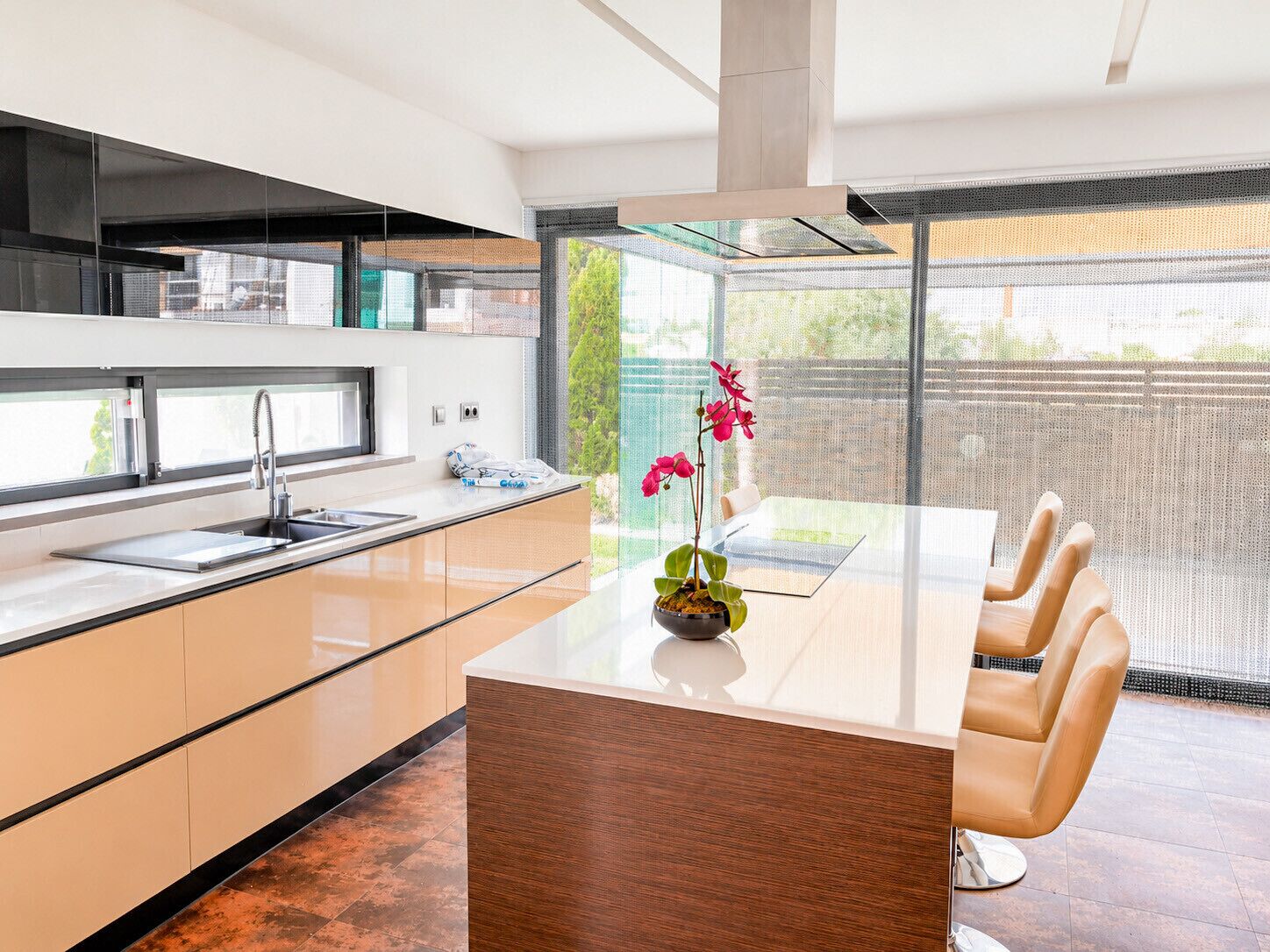 A modern kitchen with a central island, a dining area with a glass table, and a view through a large window.