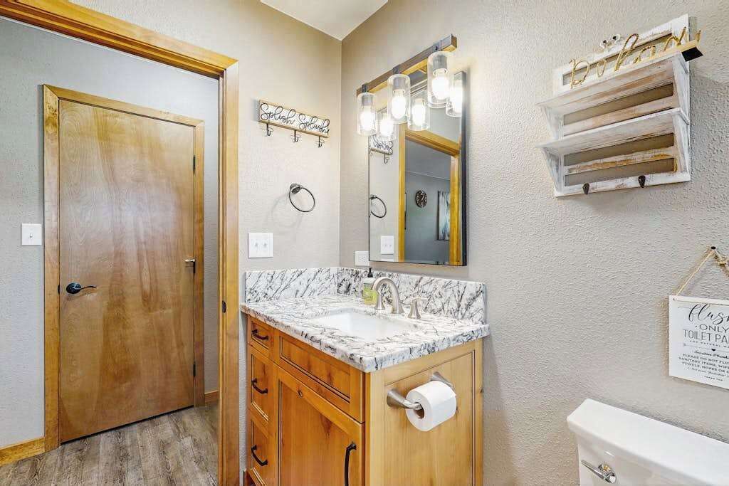 Un baño con puerta de madera, lavamanos de mármol, espejo con iluminación y un inodoro.
