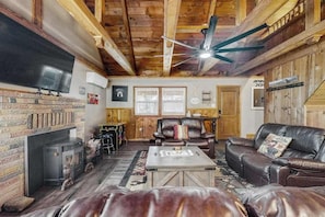 A cozy living room with a fireplace, leather sofas, a flat-screen TV, and wooden beams on the ceiling. - Forest Edge Hideaway - Musick Meadows (Shaver Lake)