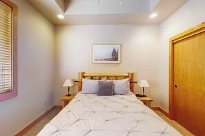 A bedroom with a wooden headboard, a bed with a white duvet, two bedside tables with lamps, a framed picture on the wall, and a wooden door. - Baby Bear Bliss - Sierra Pines (Shaver Lake)