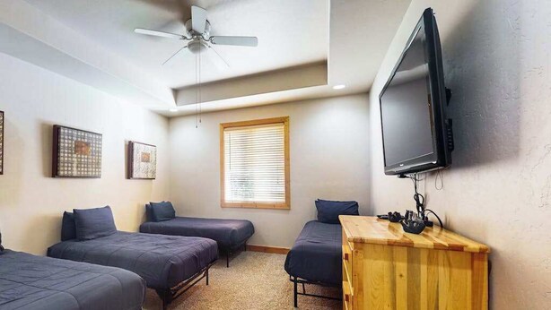 Quarto com quatro camas de solteiro, ventilador de teto, televisão presa na parede e janela com persianas.