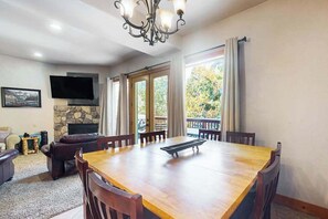 A dining area with a wooden table and chairs, a fireplace, a television, and a balcony with curtains. - Baby Bear Bliss - Sierra Pines (Shaver Lake)