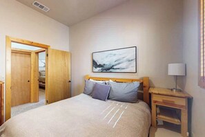 A bedroom with a bed, a bedside table, a lamp, and a door leading to another room. - Baby Bear Bliss - Sierra Pines (Shaver Lake)