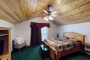 Un dormitorio con techo de madera, una cama con colcha, una silla, una mesita y una ventana con cortinas.