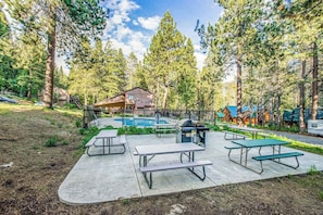 A picnic area with tables and a barbecue grill, surrounded by a pool and trees.