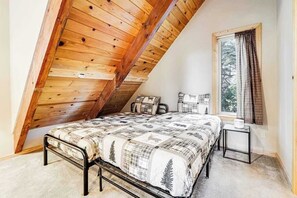 Ein Schlafzimmer mit schrägen Wänden, einem Bett mit gemustertem Überwurf, einem Fenster mit Vorhängen und einem kleinen Nachttisch.