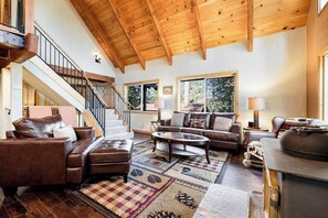 Ein geräumiges Wohnzimmer mit hoher Holzbalkendecke, einer Treppe, Ledersofas, einem Couchtisch und großen Fenstern.