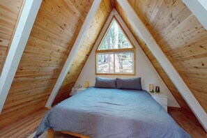 Ein raumförmiger Dachraum mit Holzbalken, einem Bett mit blauer Bettdecke, einem Fenster und einem Nachttisch mit Lampe.