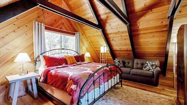 Un dormitorio acogedor con techo inclinado, una cama con estructura metálica, una mesita de noche con lámpara y un sofá con cojines.