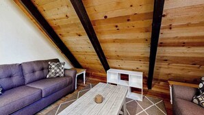 Un espacio tipo loft con techo inclinado, vigas de madera, un sofá gris, una mesita de centro blanca y un cojín decorativo estampado.