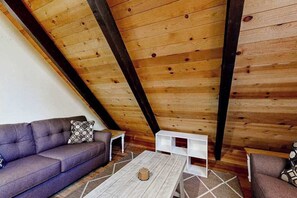 Ein Loft-Zimmer mit geneigter Decke, Holzbalken, einem grauen Sofa, einem weißen Couchtisch und einem Kissen mit Muster.