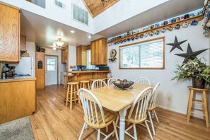 A spacious kitchen with wooden cabinets, a dining table with white chairs, and a window with blinds. - Sparrow Cove - East Village (Shaver Lake)