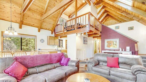 Ein großzügiger Wohnbereich mit hoher Decke und Holzbalken, ausgestattet mit zwei grauen Sofas mit roten Kissen, einem Holztisch und einem Balkon mit Geländer.