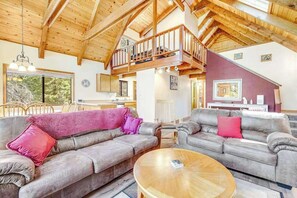 A spacious living area with a high ceiling and wooden beams, featuring two grey sofas with red cushions, a wooden coffee table, and a balcony with a railing. - Tranquility In The Woods - Blue Lake Springs (Arnold)