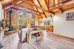 A spacious living area with a stone fireplace, wooden beams, and large windows. - Tranquility In The Woods - Blue Lake Springs (Arnold)