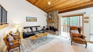 A cozy living room with a stone fireplace, a black leather sofa, a wooden coffee table, and a large window with a view of trees. - Big Mountain, Little Condo - Huntington Lake (Lakeshore)