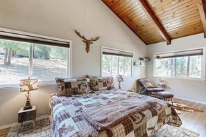 Une chambre à coucher avec un grand lit, deux fenêtres, un plafond avec des poutres en bois et une tête de cerf fixée au mur.