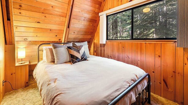 A wooden cabin bedroom with a bed, pillows, a nightstand, a lamp, and a window with a forest view. - Heavenly Daze - Meadowmont (Arnold)