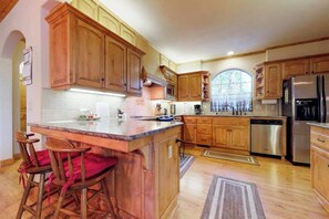 A kitchen with wooden cabinets, a stainless steel refrigerator, and a bar area with stools. - New England Cottage - Ockenden Ranch (Shaver Lake)