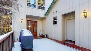 A covered outdoor area with a wooden deck, a covered barbecue grill, and a door leading to a building with a white exterior and a green roof. - New England Cottage - Ockenden Ranch (Shaver Lake)