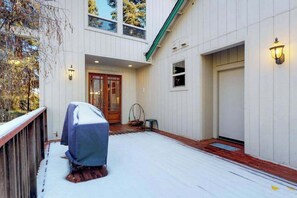 A covered outdoor area with a wooden deck, a covered barbecue grill, and a door leading to a building with a white exterior and a green roof. - New England Cottage - Ockenden Ranch (Shaver Lake)