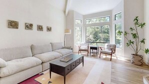 Una sala de estar moderna con un sofá, una mesa de centro y sillas, ventanas amplias y una planta en maceta.
