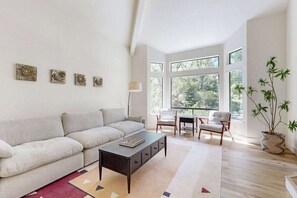 Una sala de estar moderna con un sofá, una mesa de centro y sillas, ventanas amplias y una planta en maceta.