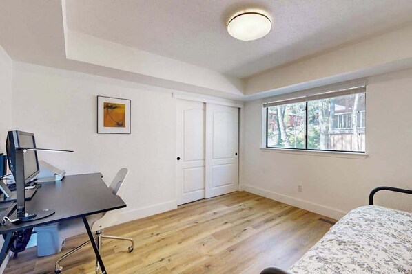A bedroom with a bed, a desk with a computer, a window, and a door.