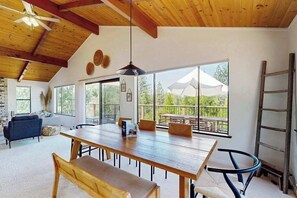 A spacious dining area with a long wooden table and benches, large windows offering a view of the outdoors, and a ceiling fan.