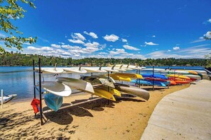 A row of canoes and kayaks lined up on a sandy beach by a calm lake.