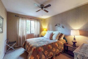 A bedroom with a bed, a ceiling fan, a window with curtains, a bedside table with a lamp, and a wall clock.