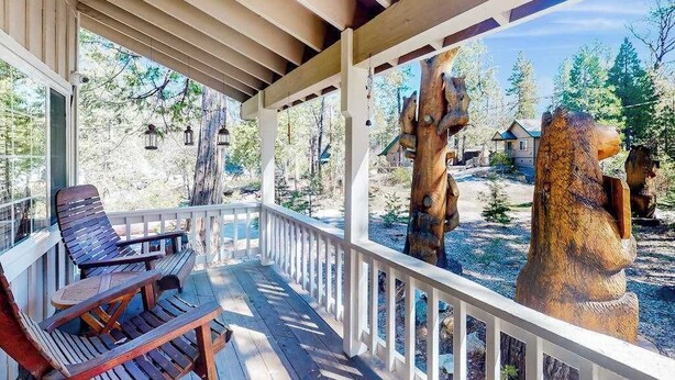A covered porch with wooden chairs and a view of a forested area with a large wooden sculpture. - Mountain Memories - Musick Meadows (Shaver Lake)