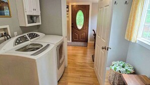 Un área de lavandería con lavadora y secadora, piso de madera, una puerta con ventanal verde y una cesta de mimbre con toallas.