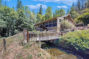 Eine Holzhütte mit einer Holzterrasse und einer Brücke über einen Bach, umgeben von Wald.