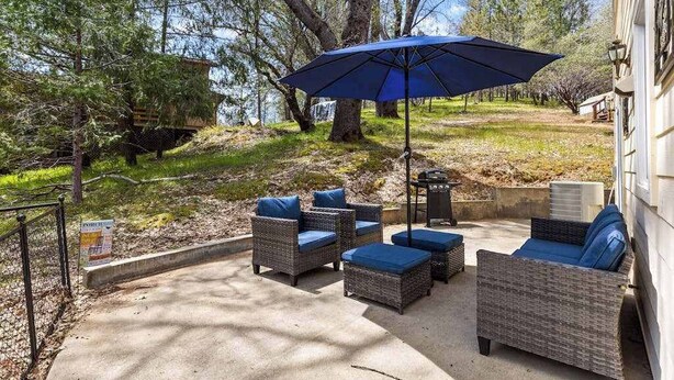 An outdoor patio with wicker furniture and a blue umbrella, surrounded by trees and a building. - Big Creek Hideaway - Pine Mountain Lake (Groveland)