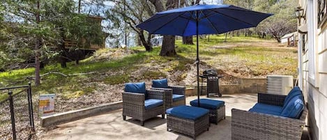 Une terrasse extérieure aménagée avec des meubles en osier et un parasol bleu, entourée d’arbres et d’un bâtiment.