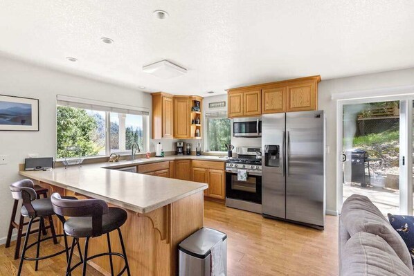 A modern kitchen with a central island, stainless steel appliances, wooden cabinets, and a large window providing a view of the outdoors. - Big Creek Hideaway - Pine Mountain Lake (Groveland)