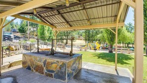 A covered outdoor area with a stone fire pit, a metal roof, and a view of a playground in the background. - Big Creek Hideaway - Pine Mountain Lake (Groveland)