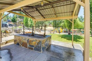 Ein überdachter Außenbereich mit einem Steinkamin, einem Metalldach und Blick auf einen Spielplatz im Hintergrund.