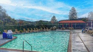 Ein rechteckiger Pool mit umlaufender Holzterrasse, Liegestühlen und einem überdachten Sitzbereich mit Tischen.