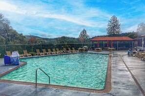 Ein rechteckiger Pool mit umlaufender Holzterrasse, Liegestühlen und einem überdachten Sitzbereich mit Tischen.