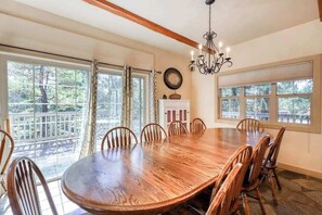 A dining room with a large wooden table surrounded by chairs, large windows with curtains, and a chandelier. - Alder Hill Hideaway - Wildflower Village (Shaver Lake)