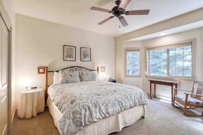 A bedroom with a large bed, a ceiling fan, two bedside tables with lamps, a rocking chair, and a window with a view of trees. - Alder Hill Hideaway - Wildflower Village (Shaver Lake)