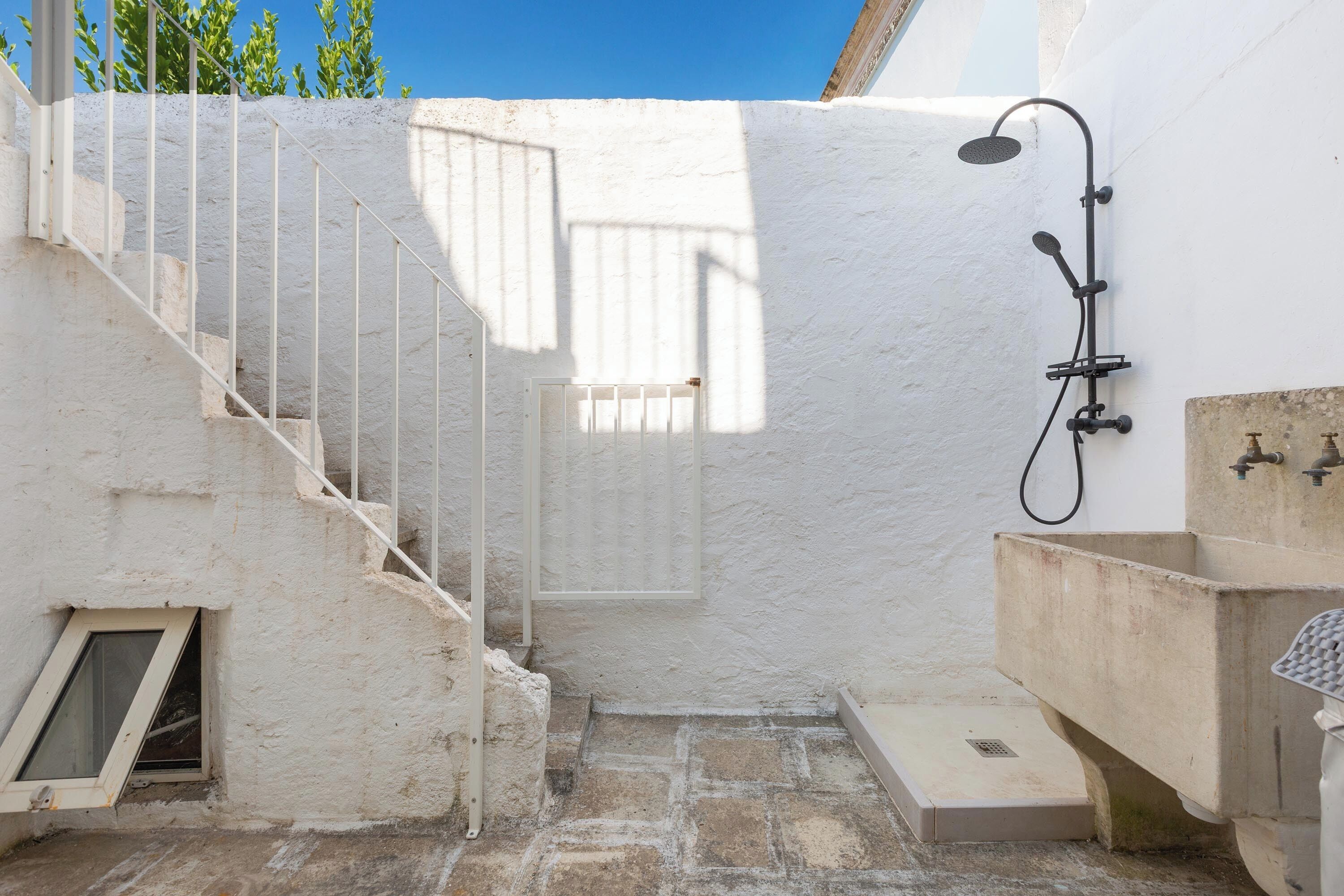 Un petit espace extérieur clos, avec un mur blanc, un escalier en métal, un espace douche et un évier.