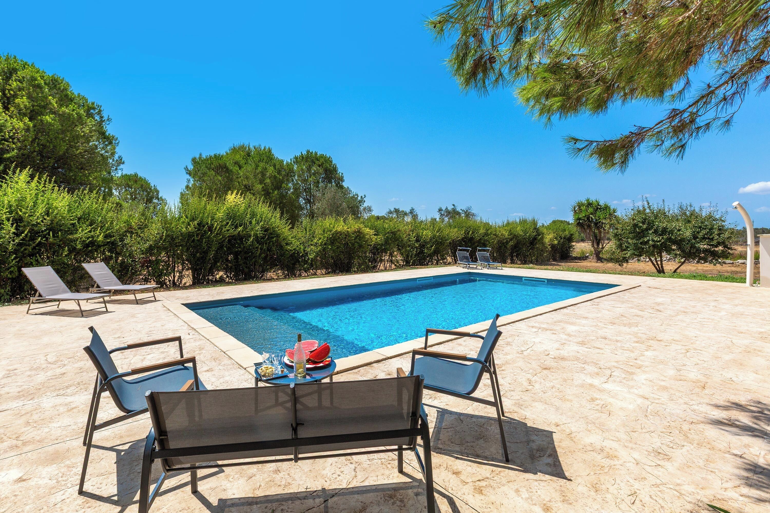 Ein rechteckiger Pool mit einer Fliesenverkleidung, umgeben von einer Sandfläche mit Liegestühlen und einem Tisch mit einer Flasche und Snacks.