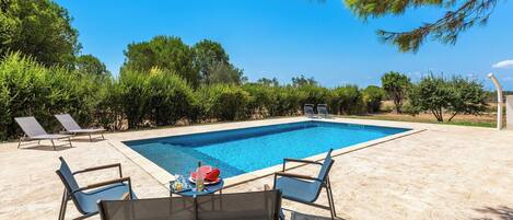 Una piscina rectangular con borde de azulejos, rodeada por una zona de arena con sillas de descanso y una mesa con una botella y aperitivos.